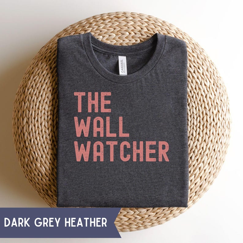 Pode incluir: T-shirt cinza escuro mesclado com o texto "THE WALL WATCHER" em coral. A camisa est&aacute; dobrada e repousa sobre uma superf&iacute;cie circular tecida. As palavras "DARK GREY HEATHER" est&atilde;o impressas em uma faixa na parte inferior.