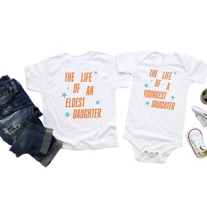 Pode incluir: T-shirts brancas e um body com o texto "THE LIFE OF AN ELDEST DAUGHTER" e "THE LIFE OF A YOUNGEST DAUGHTER" em laranja e verde-azulado. As roupas estão ao lado de um par de jeans azuis e sapatos de bebé.