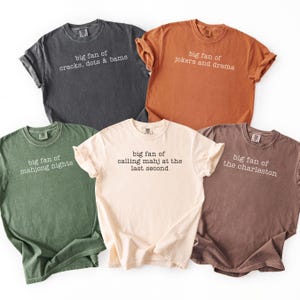 May include: Five t-shirts in various colors, including charcoal, orange, olive green, cream, and brown. Each shirt has a different phrase printed on it in white, such as "big fan of cracks, dots & bams" and "big fan of jokers and drama."
