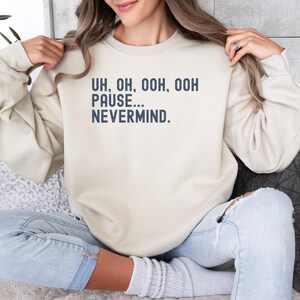 Pode incluir: Uma camisola de gola redonda bege com texto azul escuro impresso no peito que diz: UH, OH, OOH, OOH PAUSE... NEVERMIND. A peça tem mangas compridas com punhos canelados e uma gola canelada, proporcionando uma estética simples e limpa.