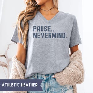 May include: A heather gray V-neck t-shirt with the text "PAUSE... NEVERMIND" in dark blue. The shirt is part of a clothing collection and is made of a soft material. The words "ATHLETIC HEATHER" are printed on a dark blue banner.
