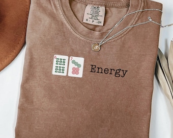 May include: A brown Comfort Colors t-shirt with the word "Energy" printed in black. The shirt features two embroidered mahjong tiles and a silver necklace with a pendant. The shirt is size small.