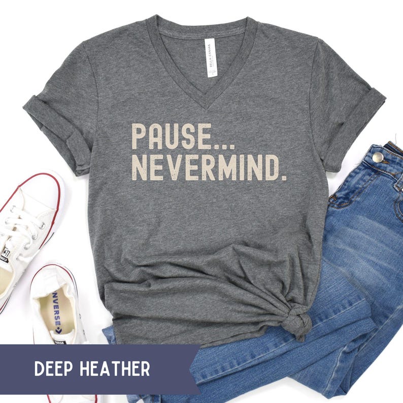 K&ouml;nnte beinhalten: Ein dunkelgraues V-Ausschnitt-T-Shirt mit dem Text "PAUSE... NEVERMIND." Das Shirt wird mit blauen Jeans und wei&szlig;en Turnschuhen kombiniert. Die Worte "DEEP HEATHER" werden auf einem dunkelblauen Banner angezeigt.