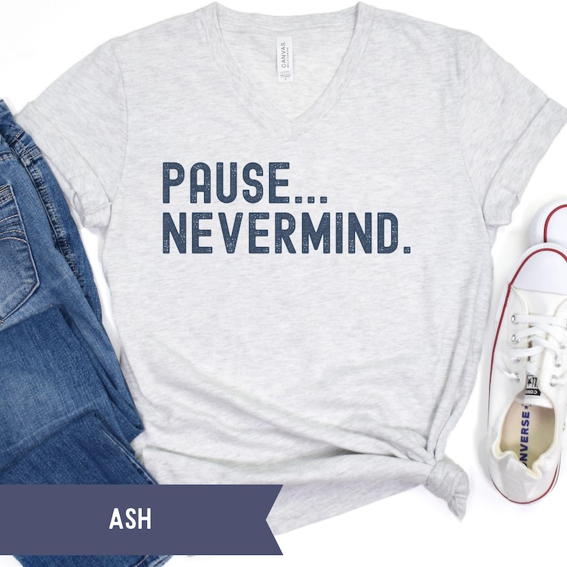 K&ouml;nnte beinhalten: Ein hellgraues V-Ausschnitt-T-Shirt mit dem marineblauen Text "PAUSE... NEVERMIND". Das Shirt wird mit blauen Jeans und wei&szlig;en Turnschuhen kombiniert. Das Wort "ASH" ist auf einem dunkelblauen Banner aufgedruckt.