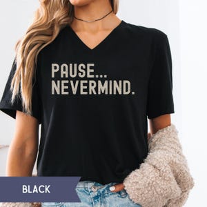 K&ouml;nnte beinhalten: Schwarzes V-Ausschnitt-T-Shirt mit dem Text "PAUSE... NEVERMIND." in einer verblassten, cremefarbenen Schrift. Das Shirt ist ein l&auml;ssiger Stil, geeignet f&uuml;r den Alltag. Das Wort "BLACK" wird in einem dunkelblauen Rechteck am unteren Bildrand angezeigt.