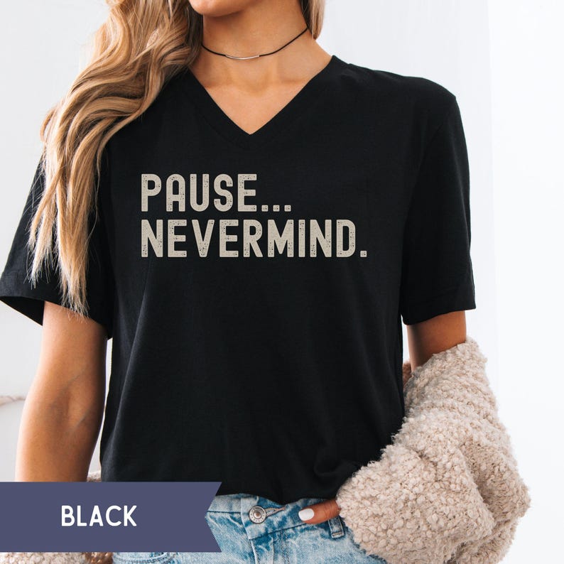 K&ouml;nnte beinhalten: Schwarzes V-Ausschnitt-T-Shirt mit dem Text "PAUSE... NEVERMIND." in einer verblassten, cremefarbenen Schrift. Das Shirt ist ein l&auml;ssiger Stil, geeignet f&uuml;r den Alltag. Das Wort "BLACK" wird in einem dunkelblauen Rechteck am unteren Bildrand angezeigt.