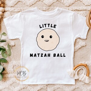 Pode incluir: Camiseta branca com uma bola de matzá de desenho animado e o texto "Little Matzah Ball".