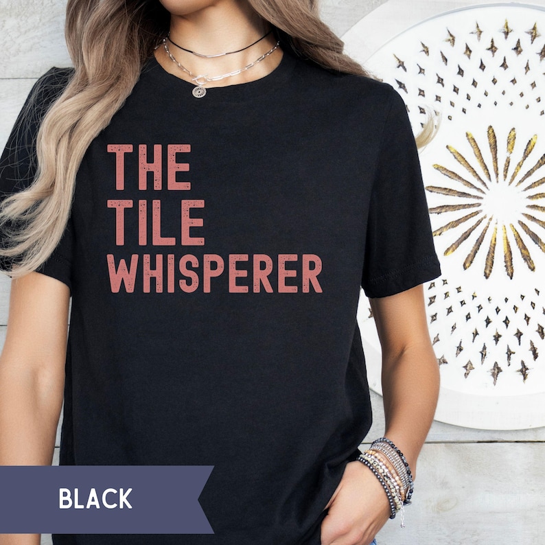 Pode incluir: Camiseta preta com o texto "THE TILE WHISPERER" em uma fonte salm&atilde;o. A palavra "BLACK" &eacute; exibida em uma faixa azul escura na parte inferior. A camiseta &eacute; de gola redonda.