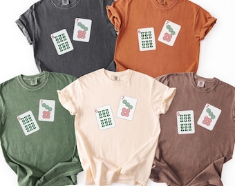 May include: Five different colored t-shirts, including gray, orange, green, cream, and brown. Each shirt features a graphic of two playing cards with the numbers six and seven. The cards have green and red details.