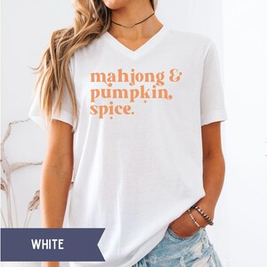 Pode incluir: Uma t-shirt branca com decote em V e o texto laranja "mahjong & pumpkin spice." com pequenos detalhes de estrelas. A peça tem mangas curtas e um corte padrão, exibindo a frase impressa claramente no centro da área do peito.