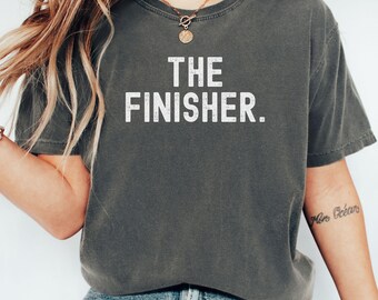 May include: Dark gray t-shirt with the text "THE FINISHER." in white block letters. The shirt is a crew neck style and appears to be made of a soft, comfortable material. The shirt is worn by a person wearing blue jeans.