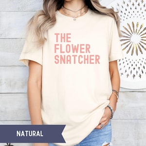 Pode incluir: T-shirt creme com o texto rosa "THE FLOWER SNATCHER". A camisa &eacute; feita de um material macio e tem mangas curtas. A palavra "NATURAL" &eacute; impressa em uma faixa azul marinho na parte inferior da imagem.