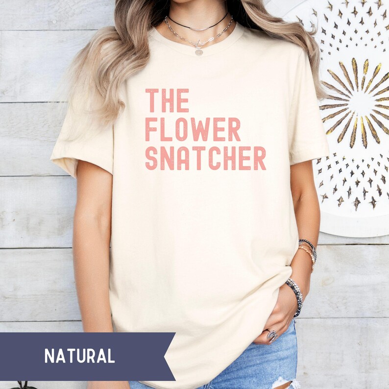 Pode incluir: T-shirt creme com o texto rosa "THE FLOWER SNATCHER". A camisa &eacute; feita de um material macio e tem mangas curtas. A palavra "NATURAL" &eacute; impressa em uma faixa azul marinho na parte inferior da imagem.