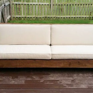 Puede incluir: Un sofá de exterior de madera con un cojín blanco. El sofá está hecho de madera marrón oscura y tiene un diseño simple y moderno.