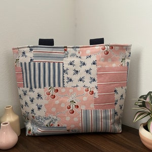 以下が含まれることがあります： ピンク、ブルー、ホワイトのストライプ、花柄、赤いチェリーが特徴の、パッチワークデザインの白いトートバッグ。バッグには黒いハンドルが付いています。