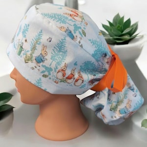 May include: A light blue scrub cap featuring a Peter Rabbit design. The cap has an orange ribbon and is decorated with illustrations of rabbits, trees, and other garden elements. The cap is on a mannequin head.