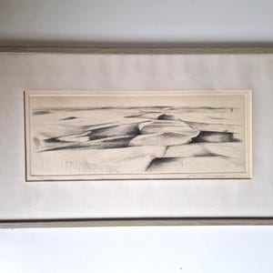 May include: A framed black and white landscape etching. The artwork shows abstract, layered forms, possibly hills or dunes, created with detailed linework. The frame is a light grey colour, and the artwork is matted.