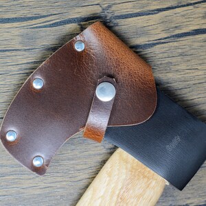 May include: A brown leather axe sheath with a black axe head and a wooden handle. The sheath has a snap closure and is secured with rivets.