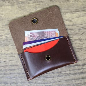 May include: A brown leather wallet with a flap that opens to reveal a red and blue card holder. The wallet has two brass snaps and is filled with credit cards and cash.