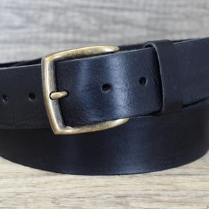 May include: A black leather belt with a rectangular brass buckle. The belt is shown coiled on a wooden surface. The buckle has a classic design, and the leather appears to be thick and durable.