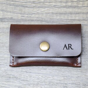 May include: A brown leather wallet with a brass button closure and the initials "AR" engraved on the front.