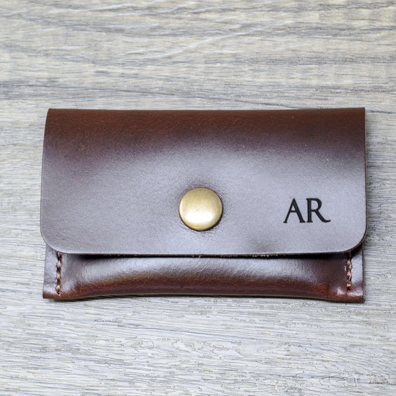 May include: A brown leather wallet with a brass button closure and the initials "AR" engraved on the front.