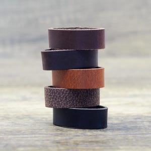 May include: A stack of five leather rings in shades of brown, from dark to black. The rings are arranged in a tower. The background is a light, neutral colour. Each ring appears to be approximately 2.5 cm in diameter.