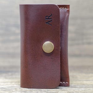 May include: A brown leather key holder with a brass button closure. The key holder has the initials "AR" embossed on the front.