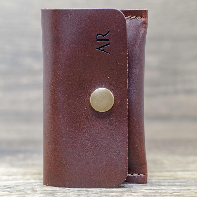 May include: A brown leather key holder with a brass button closure. The key holder has the initials "AR" embossed on the front.