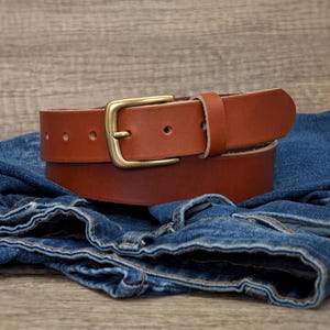 May include: A tan leather belt featuring a smooth finish and a rectangular brass-toned metal buckle. The belt includes a single leather keeper loop and multiple punched holes for adjustment, resting atop a pair of folded blue denim jeans.