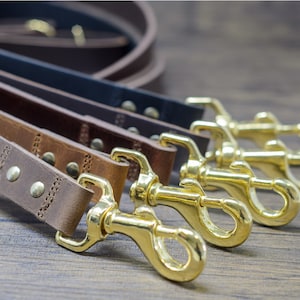 Puede incluir: Cuatro correas de cuero marrón para perros con cierres de metal dorado. Las correas están apiladas una encima de la otra, con los cierres en la parte inferior.