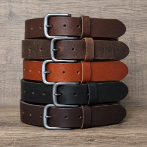 May include: A stack of five leather belts in various shades of brown and black, each featuring a matte grey metal buckle and a matching leather keeper loop. The belts are arranged vertically, showcasing different textures and leather finishes.
