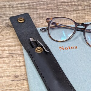 Op de afbeelding: Een zwarte leren pennenhouder met een zilveren pen erin. De houder heeft twee messing drukknopen en ligt op een blauw notitieboek met het woord "Notes" erop. Een bril met schildpadframe is ook op de foto te zien.