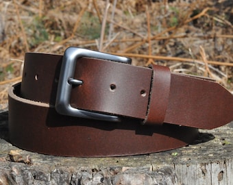 May include: A brown leather belt with a silver-colored square buckle. The belt is rolled up on a wooden surface, showcasing its rich color and texture. The belt has several holes for adjustment.