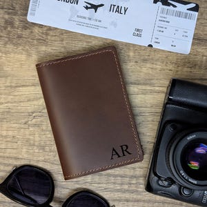 May include: A brown leather passport holder with the initials "AR" embossed on the front. The holder is lying on a wooden surface with a camera and sunglasses nearby.