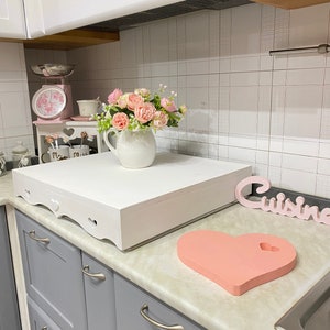 Puede incluir: Encimera de cocina blanca con una caja de madera blanca con un recorte en forma de coraz&oacute;n en la parte delantera. La caja est&aacute; sentada en la encimera junto a una tabla de cortar en forma de coraz&oacute;n rosa. Hay un jarr&oacute;n de flores rosas en la encimera detr&aacute;s de la caja. La encimera est&aacute; junto a gabinetes grises con manijas plateadas. La palabra "Cuisine" est&aacute; escrita en un letrero de madera en la encimera.