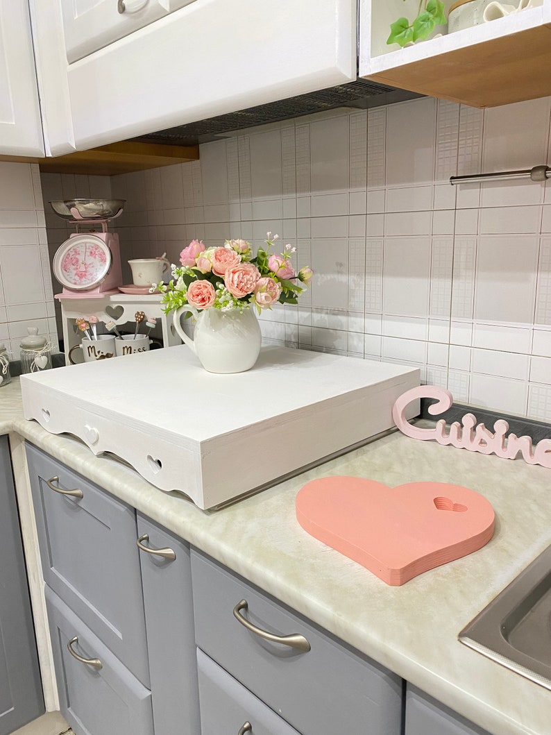 Puede incluir: Encimera de cocina blanca con una caja de madera blanca con un recorte en forma de coraz&oacute;n en la parte delantera. La caja est&aacute; sentada en la encimera junto a una tabla de cortar en forma de coraz&oacute;n rosa. Hay un jarr&oacute;n de flores rosas en la encimera detr&aacute;s de la caja. La encimera est&aacute; junto a gabinetes grises con manijas plateadas. La palabra "Cuisine" est&aacute; escrita en un letrero de madera en la encimera.