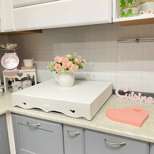 Puede incluir: Mostrador de cocina blanco con una plataforma de madera blanca con un dise&ntilde;o de corte de coraz&oacute;n. La plataforma tiene un jarr&oacute;n de cer&aacute;mica blanco con flores rosas en la parte superior. El mostrador tambi&eacute;n tiene una tabla de cortar en forma de coraz&oacute;n rosa y un letrero blanco que dice "Cuisine".