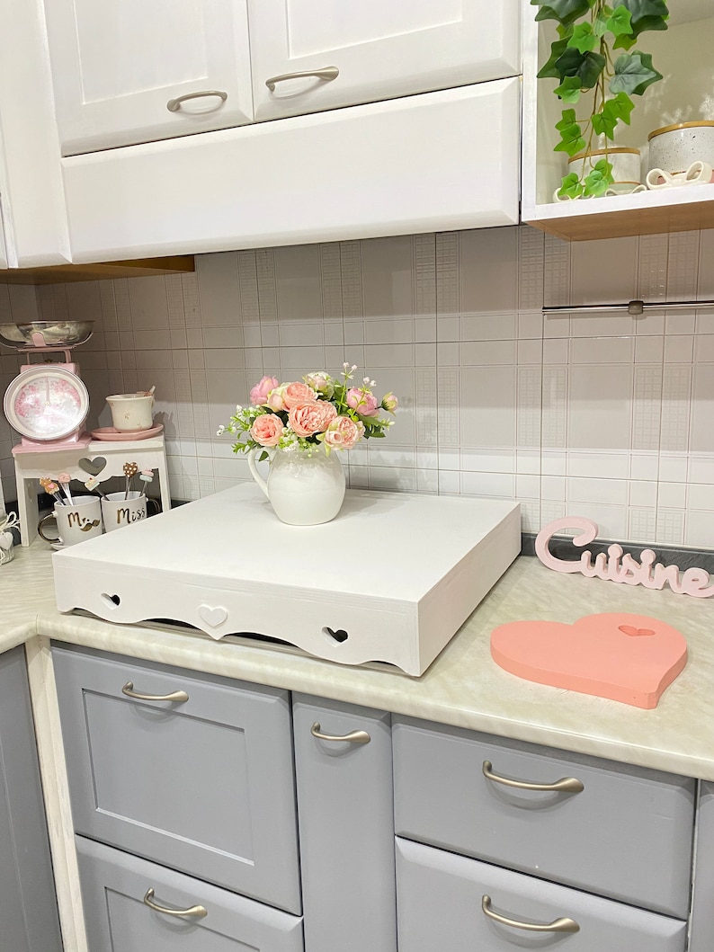 Puede incluir: Mostrador de cocina blanco con una plataforma de madera blanca con un dise&ntilde;o de corte de coraz&oacute;n. La plataforma tiene un jarr&oacute;n de cer&aacute;mica blanco con flores rosas en la parte superior. El mostrador tambi&eacute;n tiene una tabla de cortar en forma de coraz&oacute;n rosa y un letrero blanco que dice "Cuisine".
