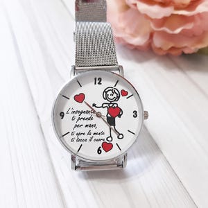 Può includere: Orologio da polso argentato con quadrante bianco con un personaggio stilizzato che tiene un cuore, circondato da cuori rossi. Il testo "L'insegnante ti prende per mano, ti apre la mente ti tocca il cuore". Cinturino in maglia d'argento.