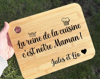 May include: A rectangular bamboo cutting board with a round handle hole. The board has black text that reads "La reine de la cuisine c'est notre Maman!" with a crown, chef's hat, and heart symbol. The text "Jules et Léo" is also present.