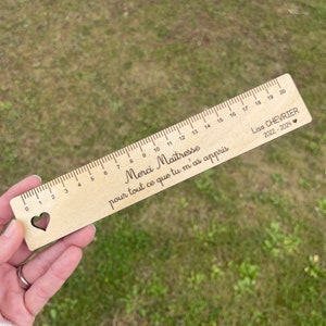 Puede incluir: Una regla de madera con un corte en forma de corazón en la marca cero. La regla está grabada con el texto "Merci Maîtresse pour tout ce que tu m'as appris" y "Lisa CHEVRIER 2022-2024".