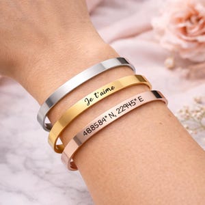 Pode incluir: Três pulseiras de punho de metal em prata, dourado e ouro rosa. A pulseira dourada tem o texto "Je t'aime" e a pulseira de ouro rosa tem as coordenadas "488584° N, 22945° E" gravadas.