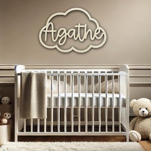 Puede incluir: Un letrero de madera en forma de nube con el nombre "Agathe" escrito en cursiva. El letrero está colgado en una pared marrón claro sobre una cuna blanca.