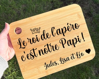 May include: A wooden cutting board with a crown and the text "Le roi de l'apéro c'est notre Papi! Jules, Lisa et Léo" in black.