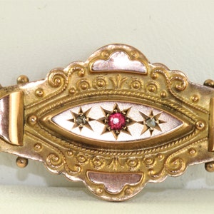 Puede incluir: Un broche de oro y oro rosa con un centro ovalado de esmalte blanco con tres estrellas con una piedra roja en la estrella central. El broche tiene un diseño ornamentado detallado.