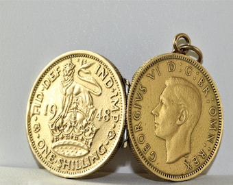 May include: A gold-colored locket featuring two detailed coin-like halves. One side displays a crowned lion and the text "ONE SHILLING", while the other shows a profile of a man and the inscription "GEORGIVS VI D.G.BR.OMN.REX".