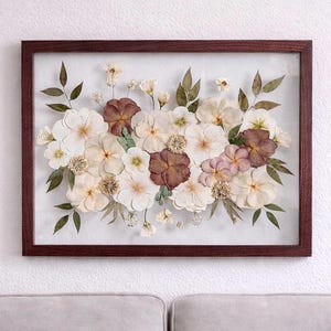May include: A rectangular wall art piece featuring a collection of pressed, dried flowers in shades of white, cream, and deep maroon, arranged with green leaves inside a dark brown wooden frame with a clear glass front, creating a botanical display.