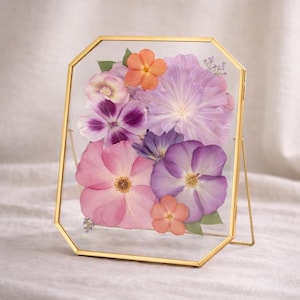 Op de afbeelding: Een achthoekige glazen lijst met een dunne gouden metalen rand met een verzameling geperste bloemen. Het arrangement bevat grote lichtpaarse bloemblaadjes, felroze bloesems en kleine oranje bloemen met groene bladeren.