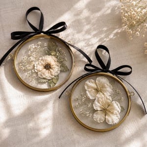 Op de afbeelding: Twee ronde messing frames met daarin geperste witte bloemen en groene bladeren achter glas. Elk frame is voorzien van een zwart satijnen lint gestrikt tot een strik, met kleine metalen ringetjes aan de zijkanten om deze decoraties op te hangen.
